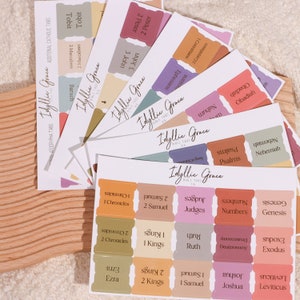 May include: A set of colorful Bible tabs with rounded corners and gold lettering. The tabs are labeled with different books of the Bible, including Genesis, Exodus, Leviticus, Numbers, Deuteronomy, Joshua, Judges, Ruth, 1 Samuel, 2 Samuel, 1 Kings, 2 Kings, 1 Chronicles, 2 Chronicles, Ezra, Nehemiah, Psalms, Obadiah, Nahum, Tobit, 1 Corinthians, Ephesians, 1 John, 2 John, 3 John, and 2 Peter.