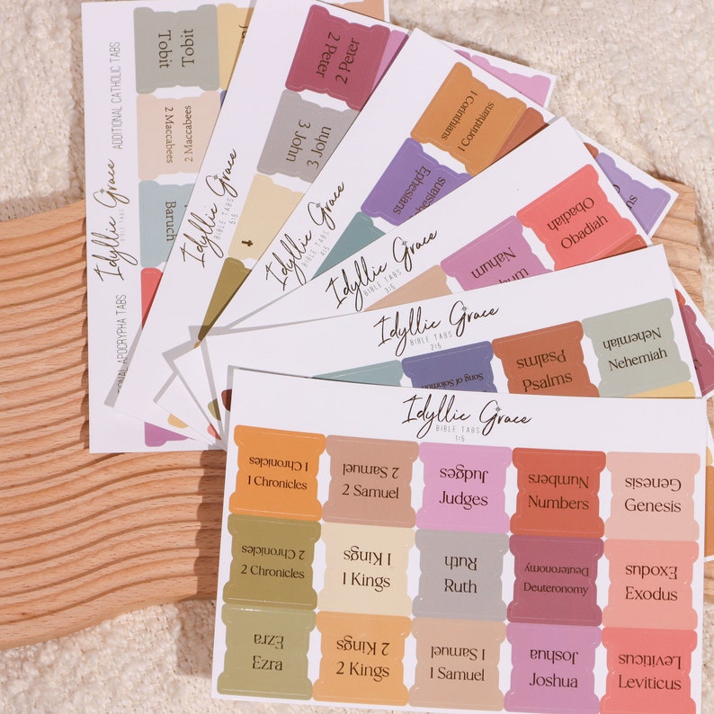 May include: A set of colorful Bible tabs with rounded corners and gold lettering. The tabs are labeled with different books of the Bible, including Genesis, Exodus, Leviticus, Numbers, Deuteronomy, Joshua, Judges, Ruth, 1 Samuel, 2 Samuel, 1 Kings, 2 Kings, 1 Chronicles, 2 Chronicles, Ezra, Nehemiah, Psalms, Obadiah, Nahum, Tobit, 1 Corinthians, Ephesians, 1 John, 2 John, 3 John, and 2 Peter.