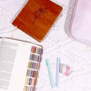 May include: An open brown leather-bound journal with colorful tabs marking different sections. The journal is open to a page with text. There are two blue and green pens and two rolls of washi tape on a white surface.