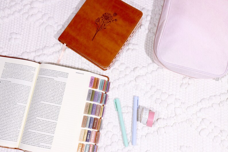 May include: An open brown leather-bound journal with colorful tabs marking different sections. The journal is open to a page with text. There are two blue and green pens and two rolls of washi tape on a white surface.
