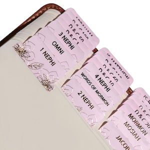 Op de afbeelding: Roze Bijbel tabs met gouden bloemaccenten en tekstlabels. De tabs zijn bevestigd aan de pagina's van een boek met een bruine leren kaft. De tabs zijn gelabeld met boeknamen en hoofdstukverwijzingen.