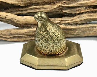 May include: A gold-toned brass figurine of a quail perched on a hexagonal base. The quail is detailed with textured feathers and a realistic beak.