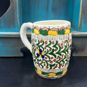 May include: A white ceramic mug with a green, yellow, red, and blue floral pattern. The mug has a handle and a rounded shape.