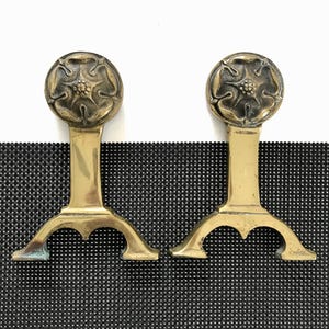 May include: Two antique brass fire dogs. Each fire dog features a floral design on top, a vertical support, and a curved base. The brass has a warm, aged patina. The fire dogs are displayed against a black and white background.
