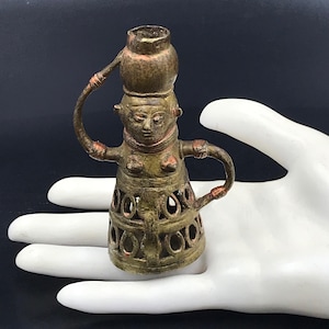 May include: A bronze figurine of a woman carrying a pot on her head. The figure has arms extended and a detailed skirt with circular cutouts. The metal has a weathered appearance with hints of red and green oxidation, suggesting age and use.