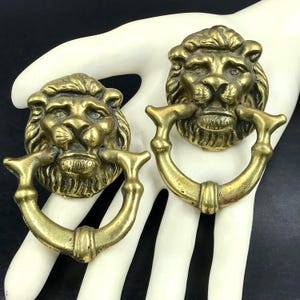May include: Two antique brass lion head drawer pulls. Each pull features a detailed lion's face with a ring handle. The brass has a warm, aged patina. These decorative pulls are suitable for furniture restoration or adding a vintage touch to cabinets.