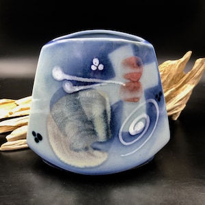 May include: A blue ceramic vase with an abstract design in white, red, and black. The vase has a rounded, rectangular shape and a smooth, glossy finish.