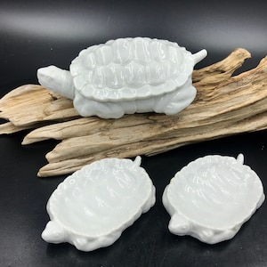May include: White ceramic turtle figurines. The image shows a large turtle figurine and two smaller turtle-shaped dishes. The turtle figurines have detailed shell patterns and are set against a dark background.