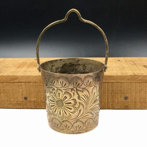 May include: A small, silver-colored metal bucket with a handle. The bucket features an engraved floral design around the body. The handle is arched and attached to the top rim. The bucket is likely vintage or antique.