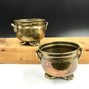 May include: Two small, round brass pots with a metallic gold finish and visible patina. Each pot features two side handles and rests on three ornate, claw-style feet. The surfaces show variations in color and texture from natural oxidation.