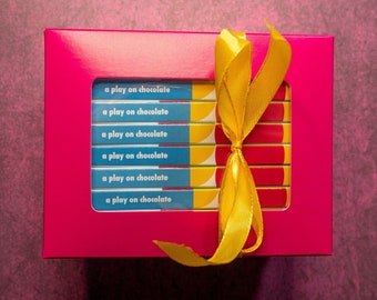 May include: A rectangular bright pink gift box with a central window displaying six stacked rectangular items. Each item features a blue label with the white text "a play on chocolate" and a yellow and red graphic. A yellow ribbon is tied around the box.