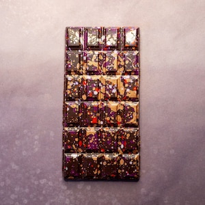 May include: A dark chocolate bar with a speckled pattern of gold, purple, and red. The bar is divided into squares.