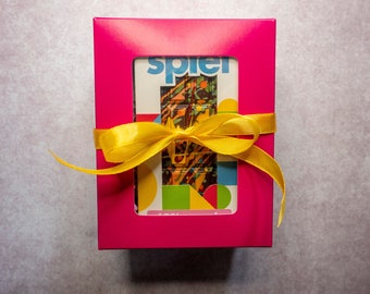 May include: A bright pink rectangular gift box with a central rectangular cutout window, tied with a yellow satin ribbon. Inside the box, a chocolate bar is visible, featuring a dark base with colorful splatters and the word spiel printed.