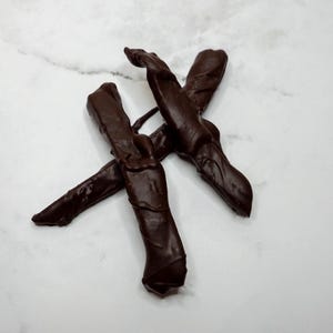 May include: Close-up of several dark chocolate-covered pretzel rods arranged on a white marble surface. The pretzels are a rich, dark brown color, with a glossy sheen from the chocolate coating. The image is well-lit, highlighting the texture of the chocolate.