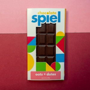 May include: A chocolate bar with the word "spiel" in blue and a geometric design. The bar is dark chocolate with the words "oats + dates" on a pink label. Net wt. 1 oz (27g).