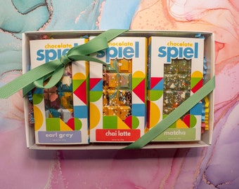 May include: A gift box with three chocolate bars tied with a green ribbon. The chocolate bars are labeled "chocolate spiel" and have different flavors: earl grey, chai latte, and matcha.