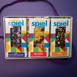 May include: A white gift box containing three chocolate bars labeled Chocolate Spiel. The bars are 78% Dominican Republic, Chai Latte, and Latte. Each bar features colorful geometric patterns on the packaging and a net weight of 1 oz (27g).