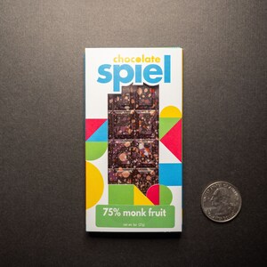 May include: A chocolate bar wrapper with a colorful geometric design. The wrapper features the text "chocolate spiel" and "75% monk fruit". The bar is a dark chocolate with a speckled appearance.