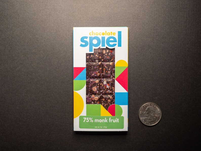 May include: A chocolate bar wrapper with a colorful geometric design. The wrapper features the text "chocolate spiel" and "75% monk fruit". The bar is a dark chocolate with a speckled appearance.