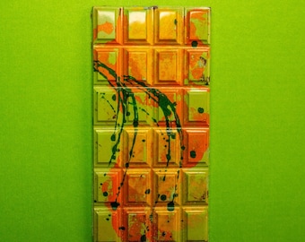 May include: A rectangular chocolate bar with a green, orange, and yellow abstract design. The chocolate is divided into squares.