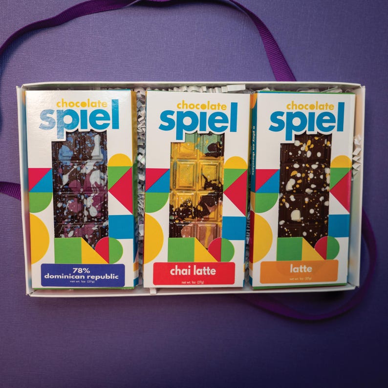 May include: A white gift box containing three chocolate bars labeled Chocolate Spiel. The bars are 78% Dominican Republic, Chai Latte, and Latte. Each bar features colorful geometric patterns on the packaging and a net weight of 1 oz (27g).
