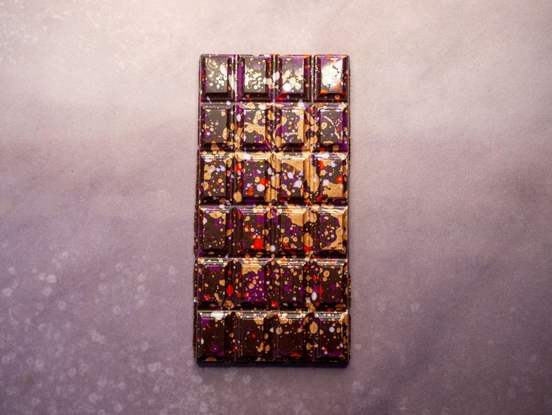 May include: A dark chocolate bar with a speckled pattern of gold, purple, and red. The bar is divided into squares.