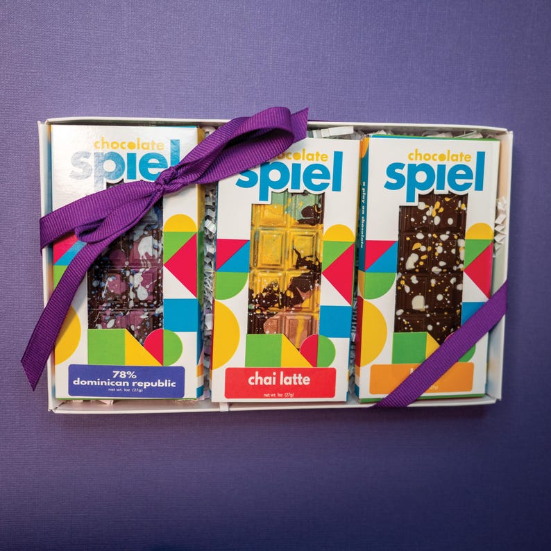 May include: A white gift box containing three chocolate bars labeled Chocolate Spiel. The left bar is 78% Dominican Republic, the center is Chai Latte, and the right is a dark chocolate bar. Each features colorful geometric packaging and a purple ribbon.