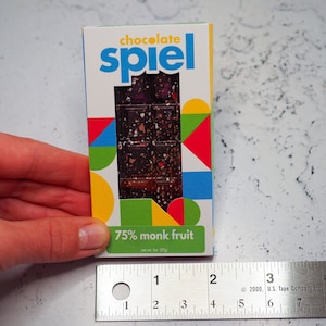 May include: A chocolate bar wrapper with a colorful geometric design. The wrapper says "chocolate spiel" and "75% monk fruit". The bar is 57g.