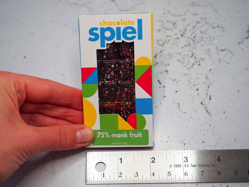 May include: A chocolate bar wrapper with a colorful geometric design. The wrapper says "chocolate spiel" and "75% monk fruit". The bar is 57g.