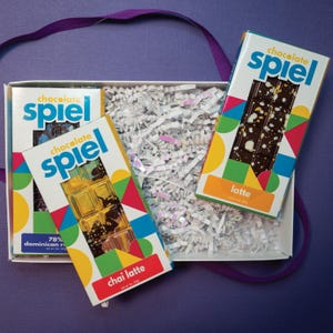 May include: A white gift box filled with shredded paper contains three chocolate bars labeled Chocolate Spiel. The bars feature colorful geometric patterns, with specific labels for Chai Latte, 78% Dominican Republic, and Latte flavors.