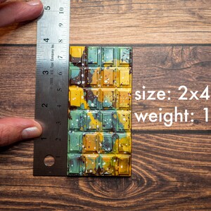 May include: A rectangular chocolate bar with a multi-colored camouflage pattern in shades of yellow, green, and brown with white paint splatters. A metal ruler shows the bar measures 2x4 inches and the text indicates a weight of 1 oz.