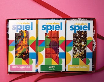 May include: A rectangular gift box containing three chocolate bars labeled Chocolate Spiel. The bars are 60% Uganda, Salty, and Peanut Butter, each featuring colorful, abstract painted designs on the chocolate surface and vibrant geometric packaging.