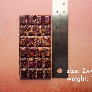 May include: A dark chocolate bar with a speckled pattern of purple, red, and gold. The bar is 2 inches by 4 inches and weighs 1 ounce.  size: 2x4 in weight: 1 oz