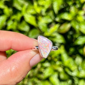 Pode incluir: Um anel de prata com uma gema de opala rosa triangular. A opala tem um acabamento brilhante e iridescente.