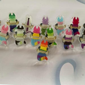 Op de afbeelding: Een verzameling heldere acrylspelden met cartoonkatkarakters in verschillende kleuren. Elke kat houdt een banner vast met de tekst "Purrride". De katten hebben regenboog-, pastel- en effen kleurenschema's.