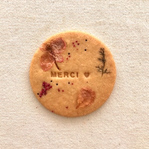 Puede incluir: Una galleta redonda de color marrón claro con la palabra "MERCI" y un corazón estampado en ella. La galleta está decorada con flores y hojas prensadas en tonos rosados y verdes.
