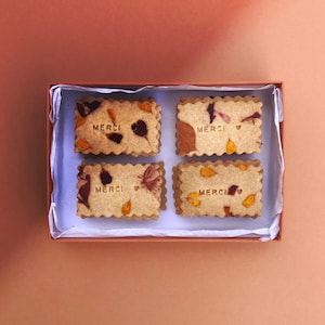 Op de afbeelding: Vier rechthoekige koekjes met het woord "MERCI" erop gedrukt. Elk koekje is versierd met geperste bloemen in tinten oranje, geel en roze. De koekjes zijn gerangschikt in een doos bekleed met wit tissuepapier.