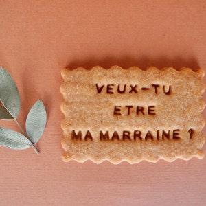 May include: A light brown cookie with the text "veux-tu etre ma marraine?" in dark brown lettering.