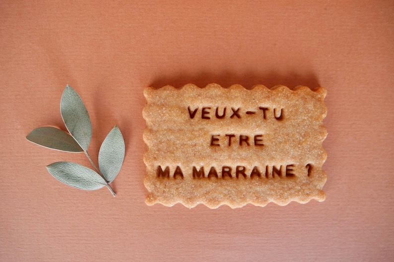 May include: A light brown cookie with the text "veux-tu etre ma marraine?" in dark brown lettering.