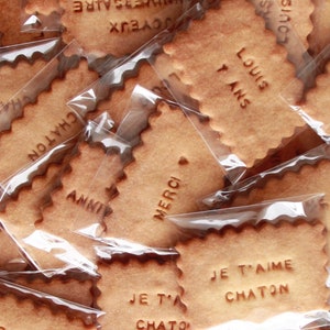 May include: A close-up of a pile of individually wrapped cookies with various French messages, including "Merci", "Je t'aime Chaton", and "Louis 7 ans".