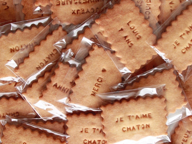 May include: A close-up of a pile of individually wrapped cookies with various French messages, including "Merci", "Je t'aime Chaton", and "Louis 7 ans".