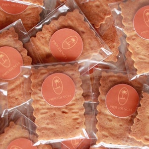 May include: A close-up of several individually packaged square-shaped cookies with a brown, textured surface. Each cookie is decorated with a round, orange sticker featuring a palm tree and the word "PROVIDENCIA".