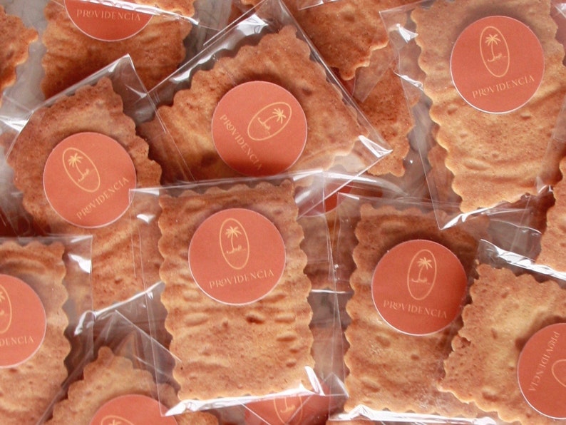 May include: A close-up of several individually packaged square-shaped cookies with a brown, textured surface. Each cookie is decorated with a round, orange sticker featuring a palm tree and the word "PROVIDENCIA".