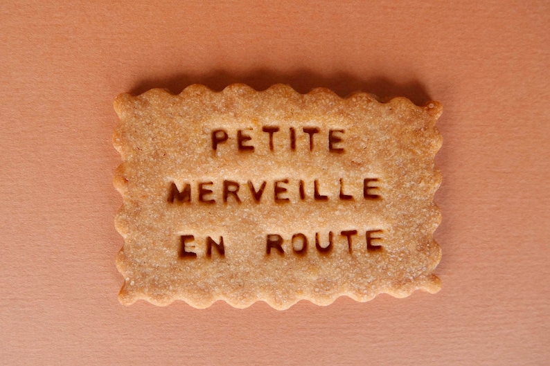 May include: A close-up of a rectangular cookie with the words "PETITE MERVEILLE EN ROUTE" embossed on the surface.