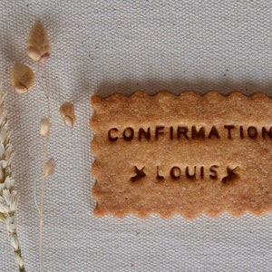 Op de afbeelding: Een bruine suikerkoekje met de woorden "CONFIRMATION" en "LOUIS" in witte glazuur.