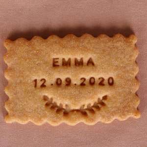 May include: A light brown cookie with the name "EMMA" and the date "12.09.2020" embossed on it. The cookie has a decorative border and a small leaf design.