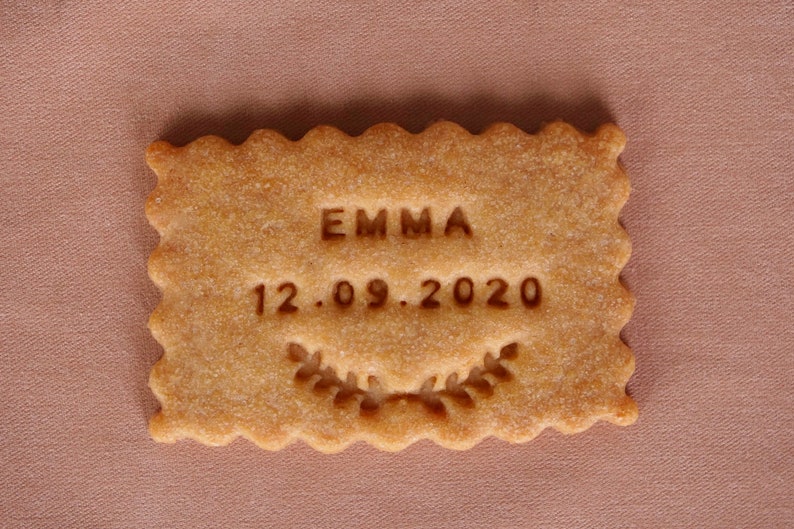 May include: A light brown cookie with the name "EMMA" and the date "12.09.2020" embossed on it. The cookie has a decorative border and a small leaf design.