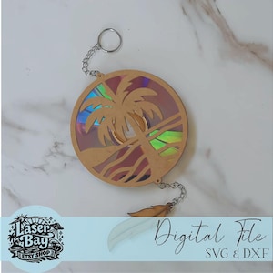 May include: A laser-cut wooden keychain featuring a tropical beach scene with a palm tree, sun, and ocean waves. The keychain has a chain and a feather charm. The text "Digital File SVG & DXF" is visible.