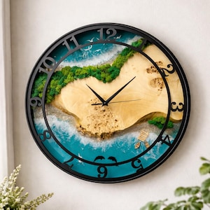 May include: A round wall clock with a black frame and a beach scene design. The clock face features a sandy beach, green island, and blue ocean. Black clock hands and numerals complete the design. The clock measures approximately 12 inches in diameter.