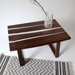 May include: A dark brown wooden coffee table with white resin inlay. The table has a curved edge and sits on a white surface with a gray and black patterned rug.